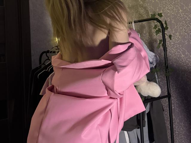 AmilySunray with blonde hair, pink off-shoulder dress, back to camera, dress falling down to reveal bare back, standing near black metal shelf with white fluffy toy.