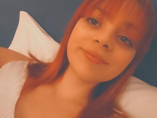 ValeryLoveLovely tiene el pelo rojo, ojos azules y una sonrisa encantadora. Lleva una camiseta blanca con textura, que deja ver sus pequeños senos, y está acostada sobre un cojín. La iluminación es cálida y suave..