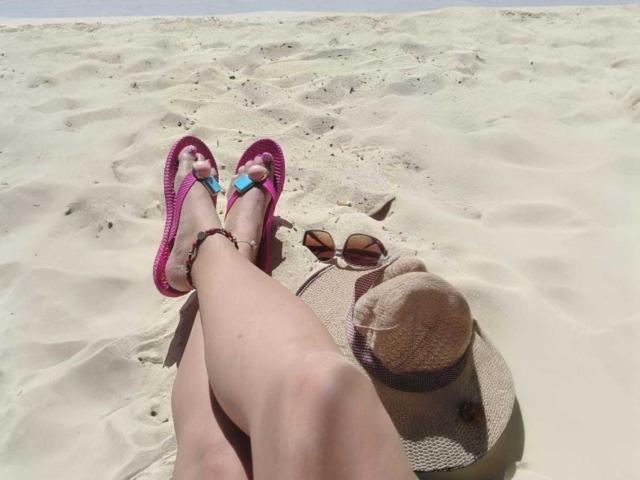 FuegoSexys Beine mit rosafarbenen Flip-Flops, blauer Fußnagellack und Armband am sandigen Strand. Sonnenhut und Sonnenbrille in der Nähe..