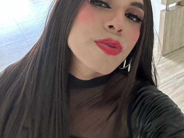 Close-up of SahraValley with long, straight black hair, wearing a sheer black top. She has pink blush on her cheeks, bold red lipstick, and black eyeliner. Her eyebrows are dark and slightly arched. She's looking slightly to the right with a subtle smile. She's wearing large silver hoop earrings. The texture of her top is smooth and slightly shiny. The background is blurred, with light-colored tiles. Her skin is fair. The image has a natural light, slightly overexposed.