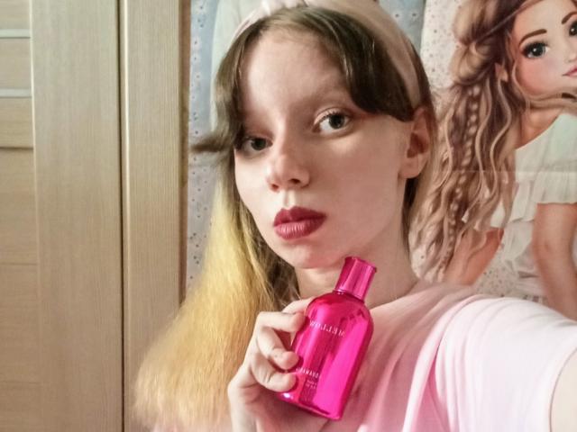KrasHulyaYourHeart met lang blond haar, roze lippenstift, die een roze fles vasthoudt, een roze shirt draagt, naar de camera kijkt en voor een pop met krullend haar staat..