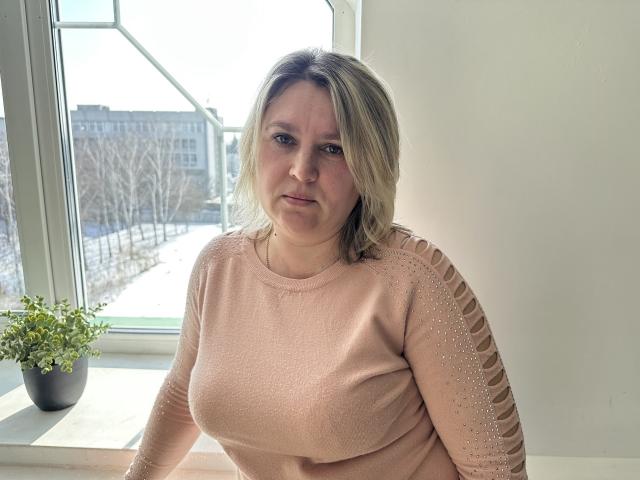 VickyRobins mit blonden Haaren, einem pinken Pullover und gepunkteten Ärmeln, sitzt am Fenster und wirkt leicht genervt. Auf dem Fensterbrett steht eine Topfpflanze. Die Brüste sind mittelgroß..