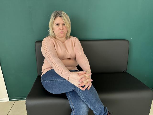 VickyRobins, avec ses cheveux blonds, son haut rose à manches longues et son jean bleu, est assise sur un canapé noir, les mains posées sur ses genoux, et a l'air légèrement contrariée. Ses seins sont petits, et ses fesses sont cachées..
