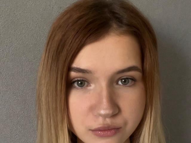RosannaHewko's face with brown and blonde ombre hair, green eyes, and pink lips, looking straight at the camera, showing a neutral expression. Her ass is not visible.