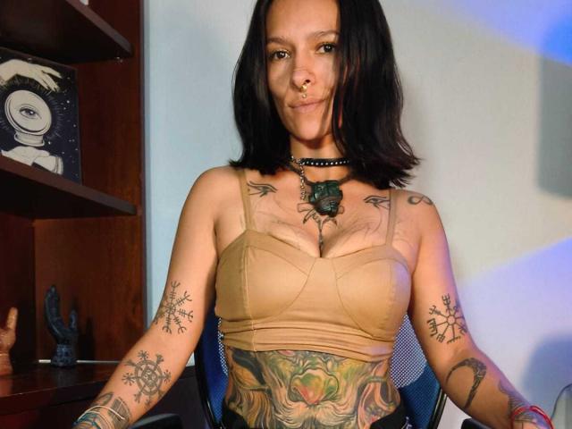 Amelie Blake, avec ses cheveux noirs mi-longs, son anneau au nez et son collier, porte un soutien-gorge beige et expose des tatouages sur ses bras, sa poitrine et le bas de son abdomen, assise sur une chaise..