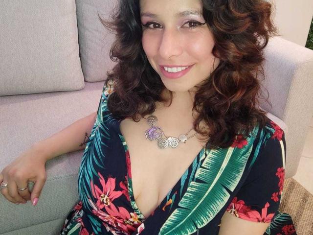 SarahWelinton with curly brown hair, smiling, wearing a floral dress with a deep cleavage, showing her tits, purple necklace, sitting on a grey couch, smiling at the camera.