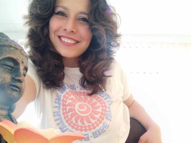 SarahWelinton with curly brown hair, smiling, wearing a white shirt with a pink mandala design, holding a book and a bronze Buddha statue in the foreground.