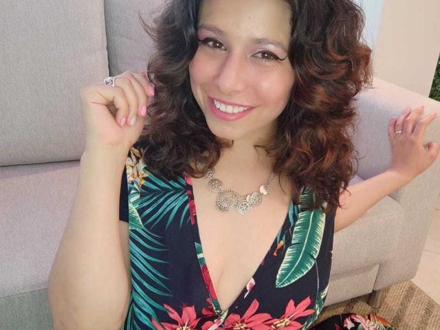 SarahWelinton with curly brown hair, smiling, pink eyeliner, and necklace, wearing a floral dress with a deep V, hands raised, showing pink nails and ring.