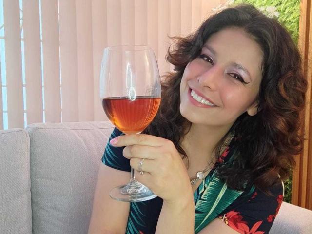 SarahWelinton with curly brown hair, smiling, holding a glass of red wine, wearing a floral dress, sitting on a gray couch, her ass slightly visible, smiling at the camera.