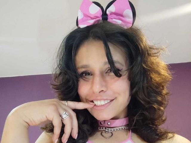 SarahWelinton with curly brown hair, pink polka-dot bow headband, smiling, pink collar with heart, touching finger to her mouth, ring on finger, cute, sexy, playful.