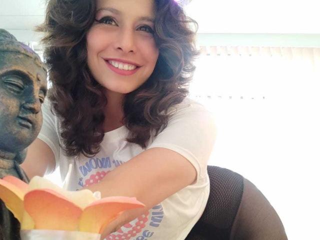 SarahWelinton with curly brown hair, white shirt, smiling, holding a flower, her ass in black mesh pants, bronze Buddha statue on the left, bright background.