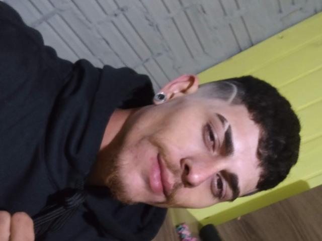 Gbsousa69, kurze, dunkle Haare mit Undercut, schwarze Kapuzenjacke, kleine schwarze Ohrstecker, leichtes Grinsen, helle Haut, sichtbarer Bartstoppeln, hält eine Kamera, gelbe Wand im Hintergrund..