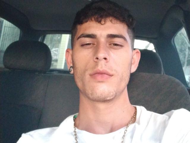 Gbsousa69 has short curly brown hair, a nose ring, small chain necklace, white shirt, and a slightly smug expression in a car. He's a young dude with a clean face.