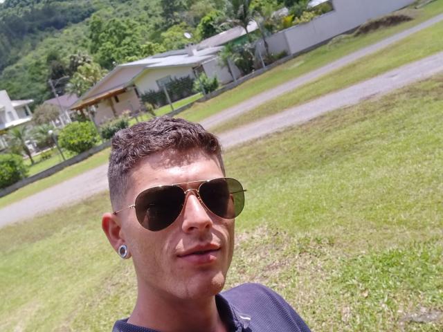 Gbsousa69, con cabello castaño corto, gafas de sol, aretes grandes y una camisa oscura, de pie en un césped, con una montaña en el fondo, mirando a la cámara con la boca ligeramente abierta..