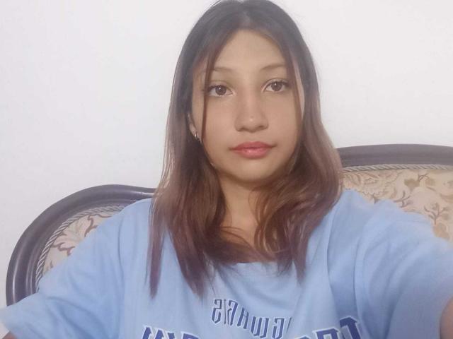 AntonelaGold has medium brown skin, straight dark brown hair, and is wearing a loose blue shirt. She's sitting with a floral upholstered chair behind her.