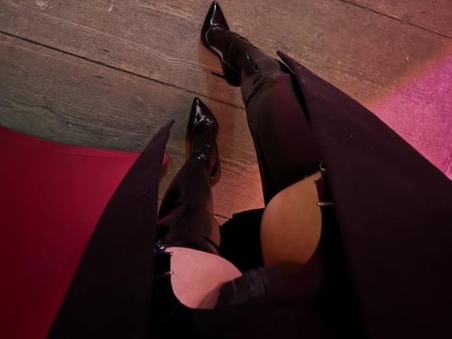 NaylaGrey's legs in black thigh-high boots with pointed toes, wearing a black skirt, sitting on a wooden floor with red and pink lighting. Boobs partially visible.