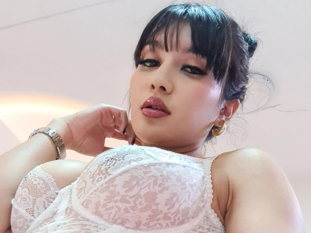 NaylaGrey with black hair in a bun, wearing a white lace bra that shows her big tits. She's looking down, her pink lips slightly parted, and has a silver watch.