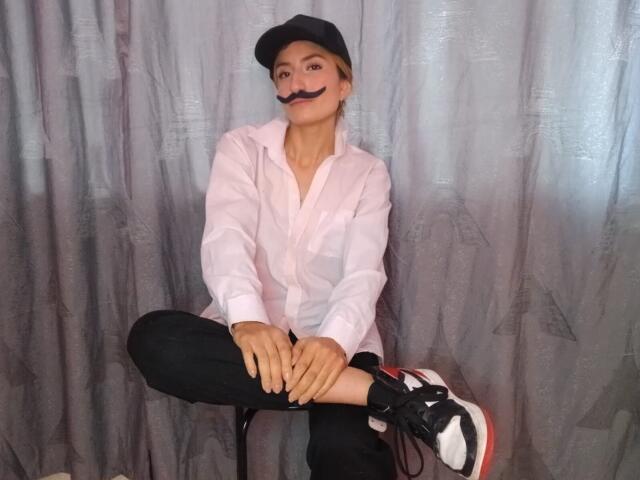 Kaiix sits cross-legged against a grey curtain. She wears a black cap, white button-up shirt, and black pants. A black mustache is digitally added to her face. She has light skin and blonde hair. She wears black and white sneakers with red soles. Her hands rest on her knee. The image has a casual, humorous style. The focus is on Kaiix's quirky outfit and the digital mustache. The textures are smooth with the curtain's subtle pattern.