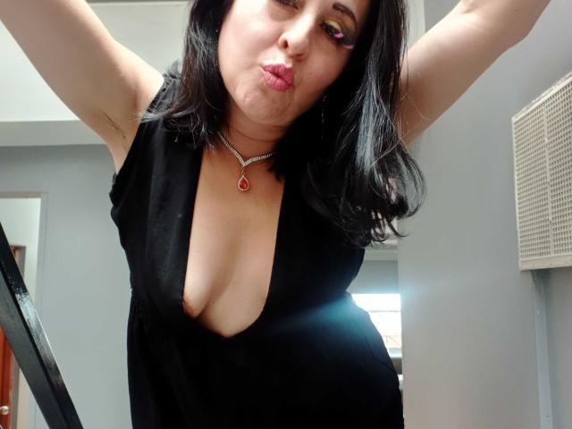 ScarletMilfX, a medium-skinned woman with black shoulder-length hair, pouts her lips. She wears a deep black dress with a plunging neckline, exposing her cleavage. She has pink eyeshadow and a red teardrop necklace. Her arms are raised, and she's taking a selfie from a low angle. The image focuses on her upper body and face. The background is a simple room with a door and vent. The photo is candid and slightly angled.