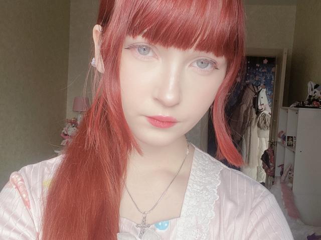 EilenePulford with red hair, bangs, pale skin, blue eyes, pink lips, white lace top, silver necklace, looking at camera, in well-lit room, relaxed expression, long side ponytail.