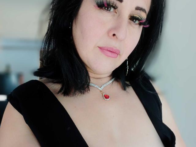 Close-up of ScarletMilfX. She has pale skin, black shoulder-length hair, and pink lipstick. Her eyes have heavy, colorful eye makeup with pink and green shades. She wears a black sleeveless top with a deep neckline, revealing a small amount of cleavage. A silver necklace with a red gemstone pendant rests on her chest. Her expression is neutral, slightly looking to the right. The background is blurred. The image has a soft, natural light. Her makeup and jewelry are the focal points.