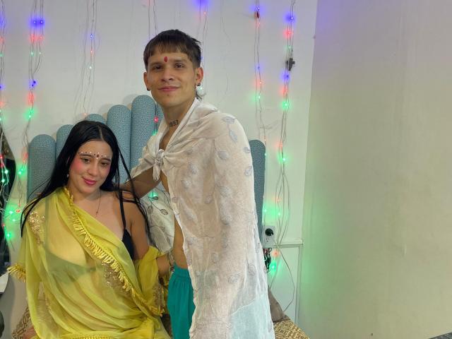 AnjaliRohan with black hair in a yellow saree and AnjaliRohan with brown hair in a sheer white top, standing together, colorful lights in the background. Her tits are visible.