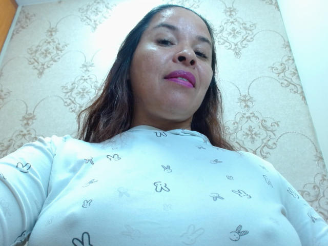 Low-angle selfie of LizethBells with light brown skin and long, wavy brown hair. She wears a white, long-sleeved top with small, black butterfly patterns. Her pink lipstick stands out against her slightly shiny skin. The background features beige, floral-patterned wallpaper. The light source is above and to the right, creating a slight shadow on her face. The perspective emphasizes her chest and facial features.