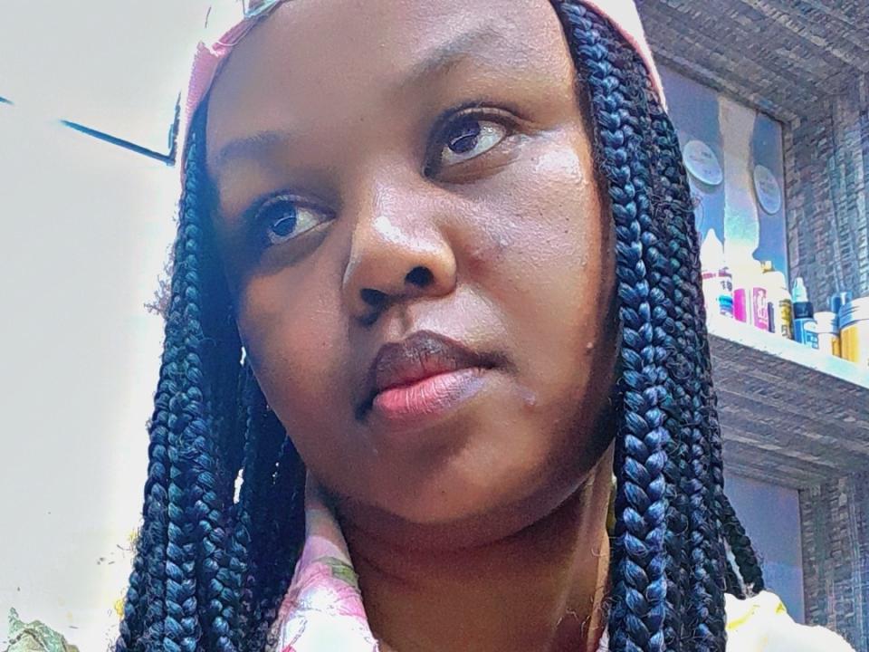 Raiy tiene la piel de color marrón oscuro, trenzas negras largas y un pañuelo rosa en la cabeza, mirando ligeramente hacia un lado con una sonrisa sutil. Sus labios están ligeramente entreabiertos, mostrando una expresión relajada..