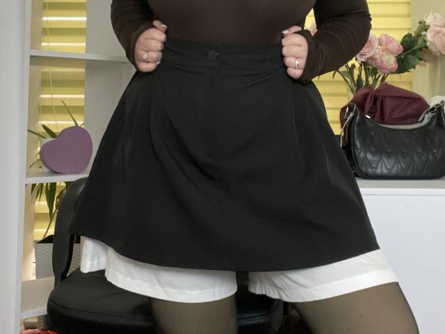 Das braune Oberteil von Madrinia, der schwarze Rock, das weiße Hemd darunter und die schwarzen Strumpfhosen, Hände, die den Rock hochziehen und ein wenig des weißen Hemdes zeigen. Schwarze Handtasche, rosa Rosen, Hintergrund unscharf..