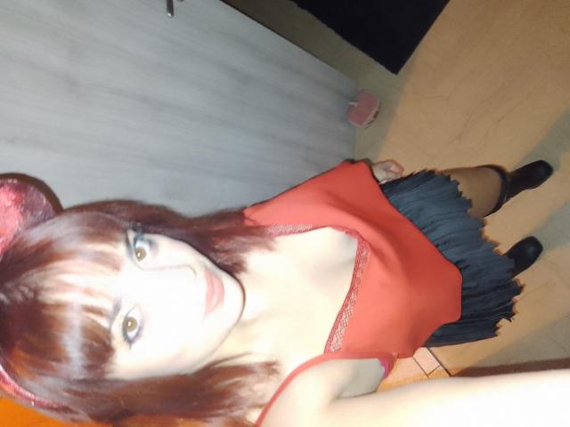 ValeryLoveLovely, con cabello rojo, flequillo y una camiseta roja que deja ver sus pechos, vestida con una falda azul y botas negras, de pie en una habitación con una puerta de madera..