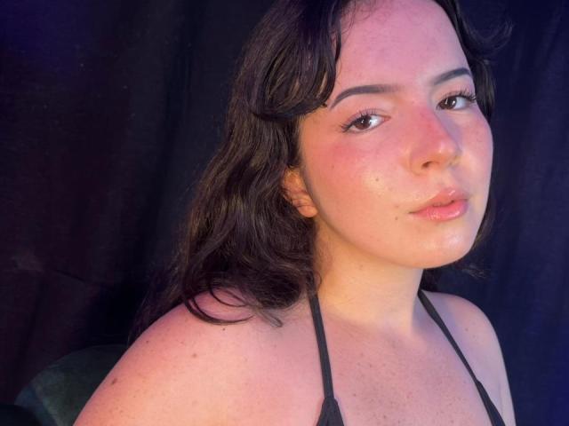 LiliBonnet has fair skin, black hair, and subtle makeup with pinkish cheeks, wearing a black halter top, showing her tits and a cute, slightly flushed face.
