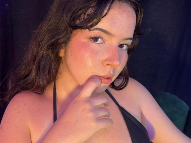 LiliBonnet with freckled face, dark wavy hair, and pink cheeks, biting her finger, wearing a black halter top, showing her small tits.