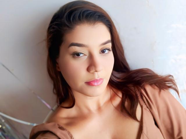 Paulalivia, con piel de color marrón claro, cabello marrón oscuro y lápiz labial rosa, mirando a la cámara con una sonrisa sutil, vestida con una camisa color beige, mostrando sus hombros desnudos..