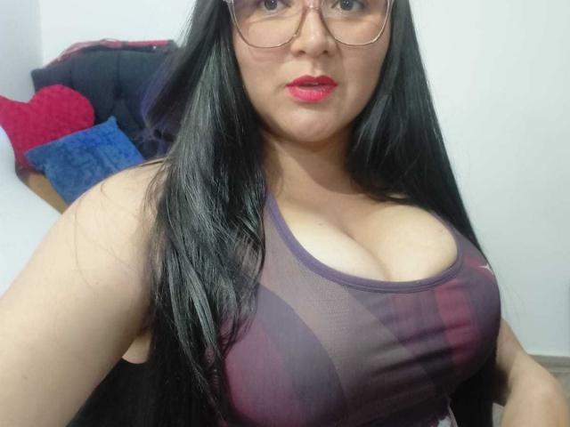 Philipa with long black hair, red lipstick, glasses, and a sheer purple sports bra showing her large tits. She's sitting with a slightly serious expression.