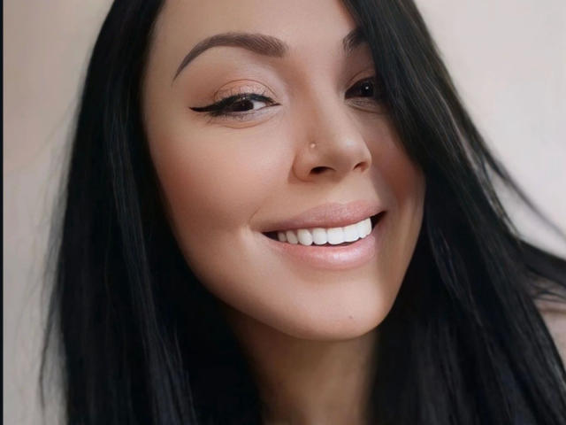 Close-up of Layla8-hot with long black hair, light brown skin, and a subtle smile. She has well-defined eyebrows, black winged eyeliner, and a small nose stud in her right nostril. Her white teeth contrast with her light pink lips. The image has soft lighting, highlighting her smooth skin and natural beauty. The composition focuses on her face, with her hair slightly parted. The overall style is clean and modern.