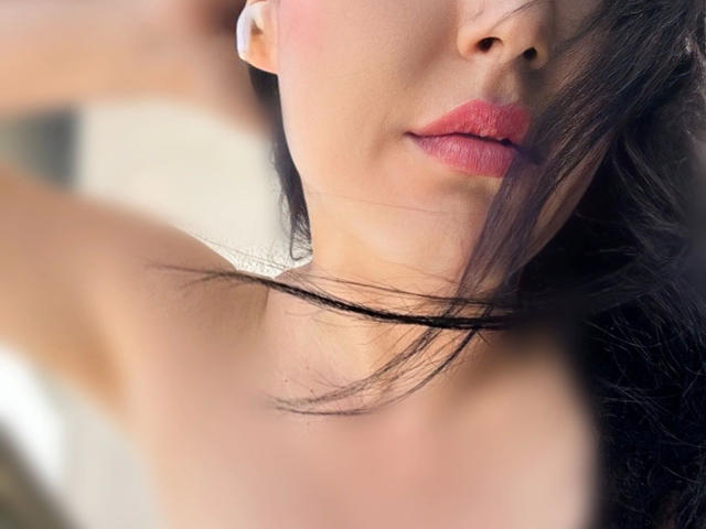 Layla84-hot's face, showing pink lipstick, slightly parted pink lips, black hair, and a fair skin tone, with one arm raised, highlighting her bare shoulder.