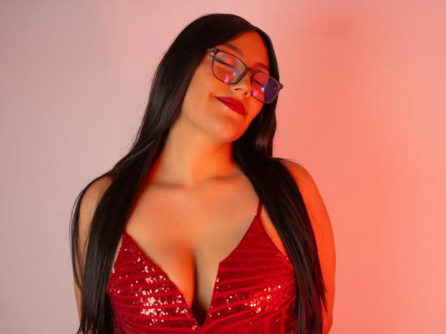 IsabellCollin with long black hair, red sequined dress, cleavage, and glasses, smiling with eyes closed, red light on right side, showing her tits.