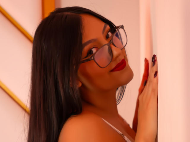 IsabellCollin with long black hair, red lipstick, and glasses, leaning against a wall, showing off her red nails and smiling with her head tilted.