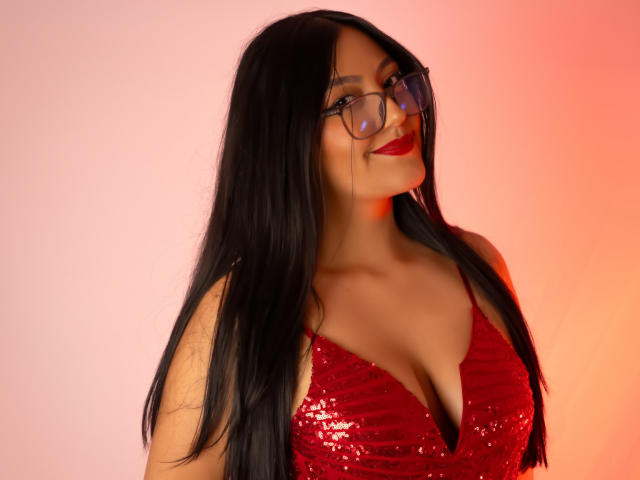 IsabellCollin with long black hair, wearing red sequined dress, glasses, and red lipstick, smiling, showing her big tits, against a pink and orange gradient background.