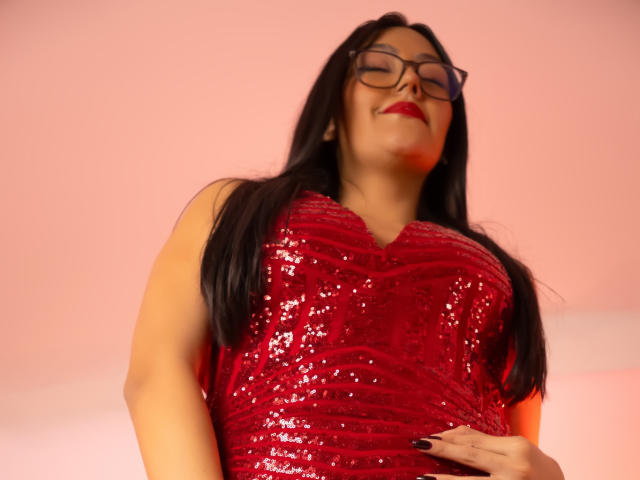 IsabellCollin in a red sequined dress, her large tits prominent, long black hair, glasses, red lipstick, and black nails, eyes closed, looking down with a slight smile.