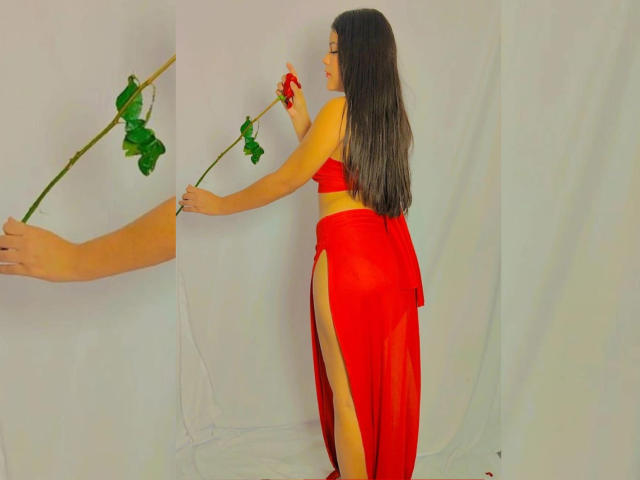 BadKristty with long dark hair, wearing a red sheer dress with high slits, holding a rose, her ass and back visible, standing next to a hand holding another rose.