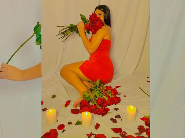 BadKristty in a red dress, kneeling, holding roses, covering face, surrounded by candles, rose petals on white cloth, right hand holding rose, dark hair, soft lighting.