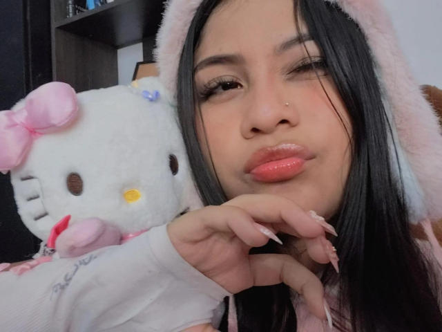 BadKristty with black hair, pink hood, and glossy lips, winking, holding a white Hello Kitty stuffed toy, pink bow, and pink nails, in a selfie.