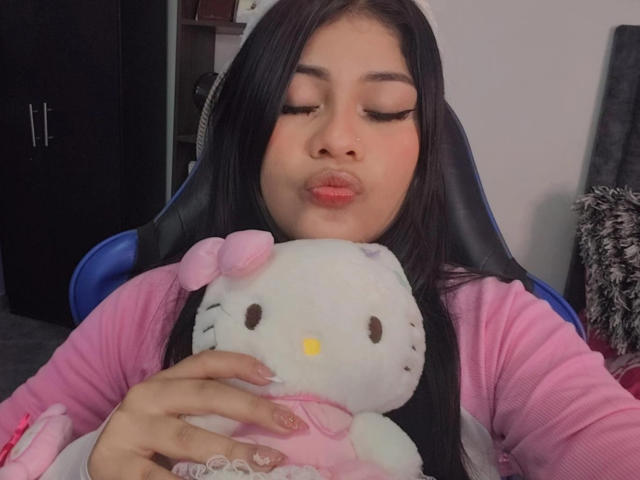 BadKristty with long black hair, pink lipstick, and closed eyes, kissing a white Hello Kitty stuffed toy, wearing a pink shirt, sitting in a blue chair.
