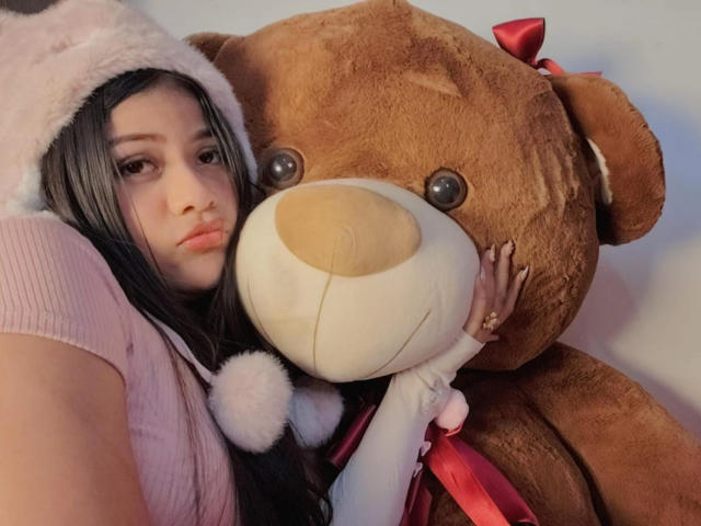 BadKristty with long black hair, pink top, and white fuzzy hat, hugging a big brown teddy bear with a red ribbon, holding its face. Her eyes are half-closed.