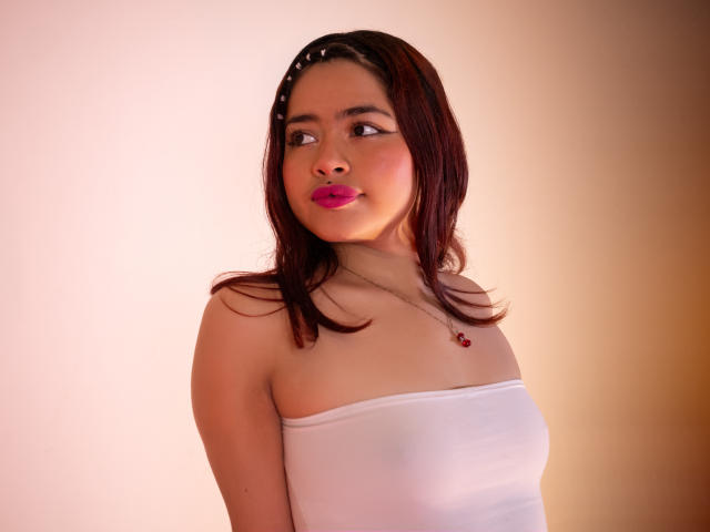 JennieRider with medium brown skin, pink lipstick, dark red hair, white strapless top, small pink necklace, looking to the side, soft light, subtle smile, sexy, cute.