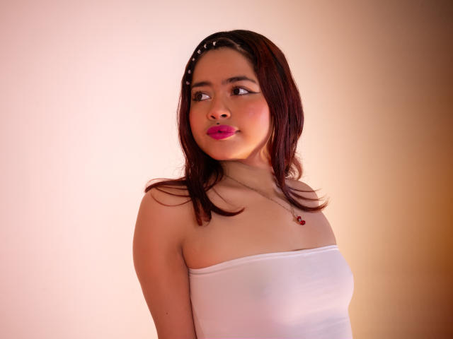 JennieRider with pink lips, brown hair, white strapless top, small tits, necklace with red pendant, looking to the side, soft light, slight smile, headband with small white dots.