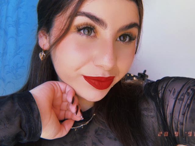 VenusxMusa with red lipstick, black sheer top, long brown hair, green eyes, and red lipstick, holding her chin, wearing gold earrings, looking at the camera.