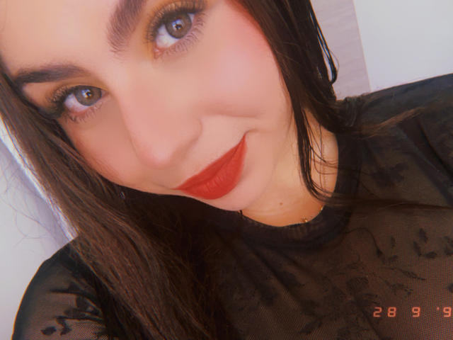 VenusxMusa mit rotem Lippenstift, einem schwarzen, transparenten Oberteil, braunen Augen und langen, dunklen Haaren, lächelt in die Kamera. Die Brust ist nicht sichtbar, der Fokus liegt auf ihrem Gesicht..