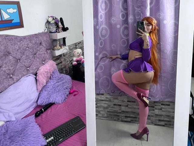 MelannyTaylorr with long orange hair, purple crop top, pink thigh-highs, and purple heels, showing her big ass and small tits in a mirror selfie, standing on one leg.